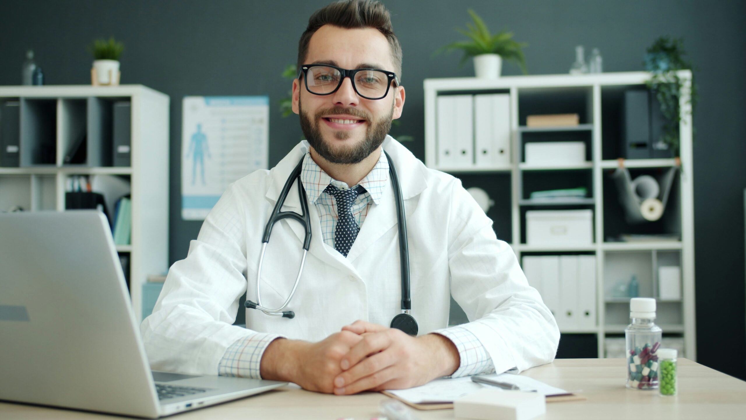 A young male doctor wearing glasses sits at a desk with a laptop