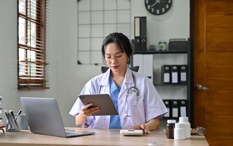 A doctor wearing a white lab coat and a stethoscope using a calculator and holding a tablet